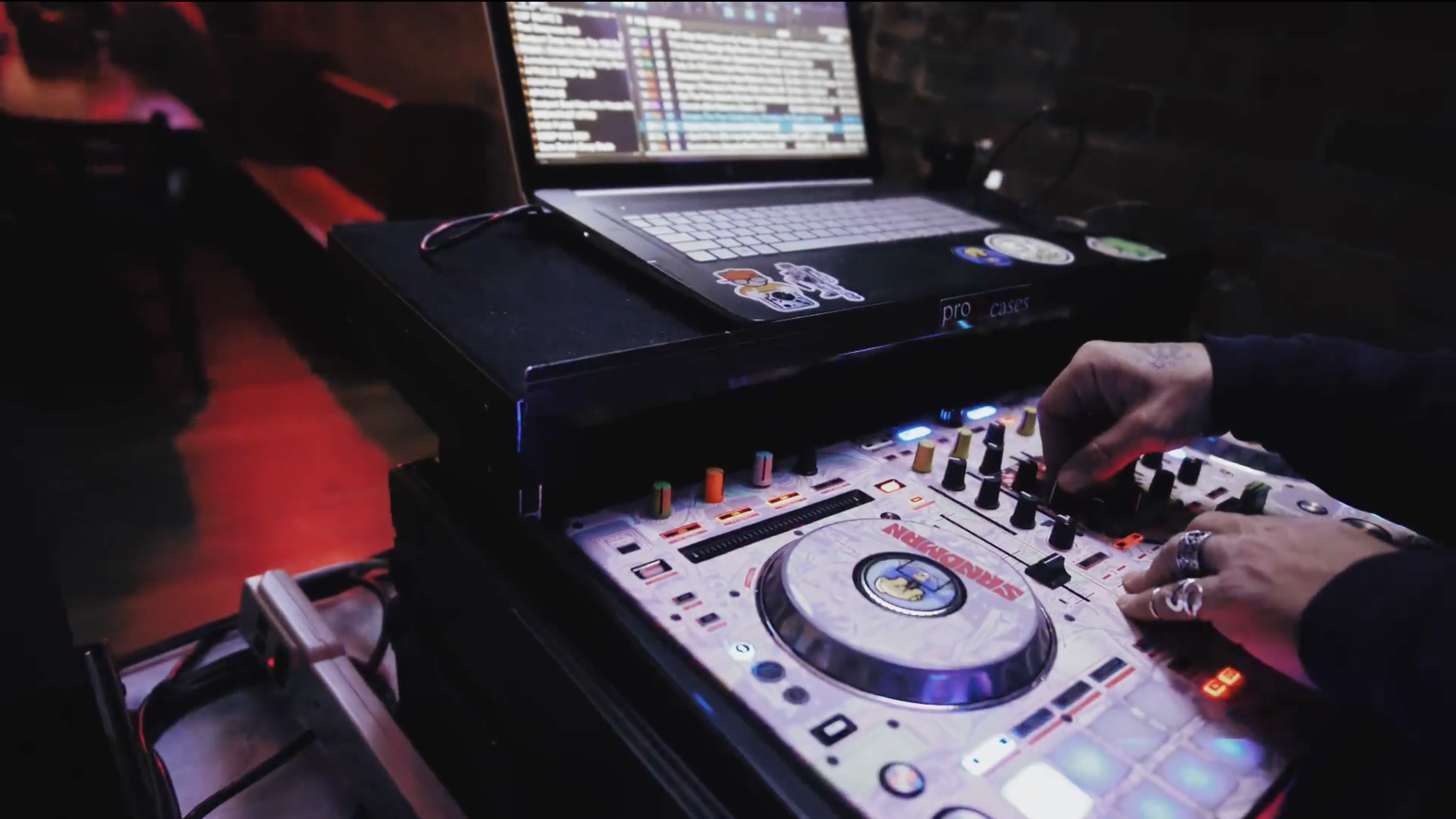 A pair of hands sporting silver rings turn nobs on a turntable. Above the turntable sits a laptop.