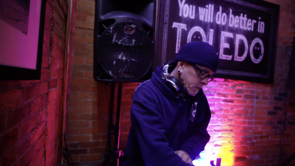 A person with a goatee wearing a black beanie and black hoodie looks intently down at something out of frame. There are headphones around their neck and there are colorful pink and yellow lights and a large sign hanging on the brick wall behind them that reads "You Will Do Better in Toledo."