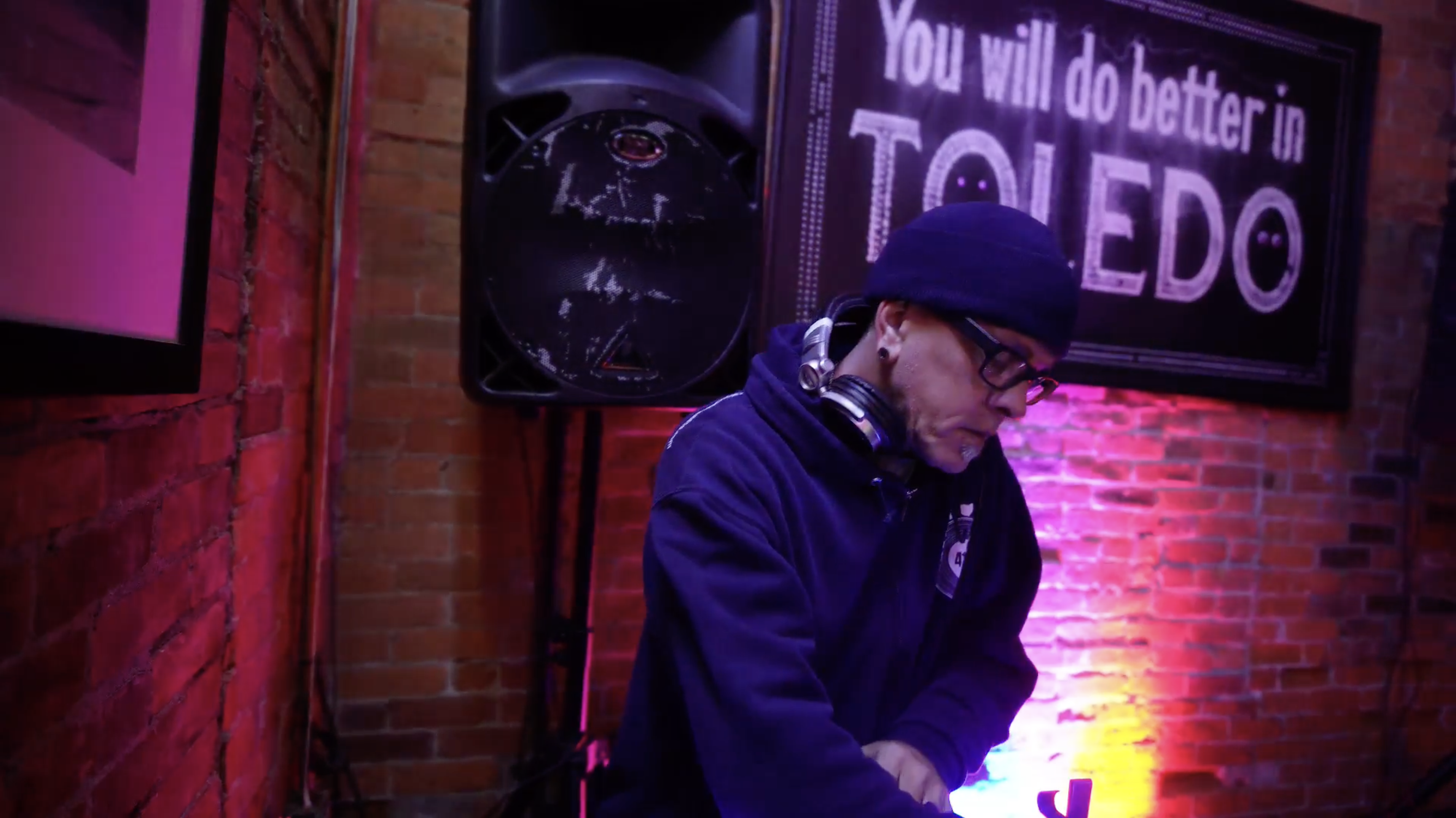 A person with a goatee wearing a black beanie and black hoodie looks intently down at something out of frame. There are headphones around their neck and there are colorful pink and yellow lights and a large sign hanging on the brick wall behind them that reads "You Will Do Better in Toledo."