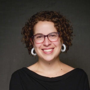 Headshot of a smiling person of light skin tone, with brown short curly hair, and wearing a dark shirt and hoop earings