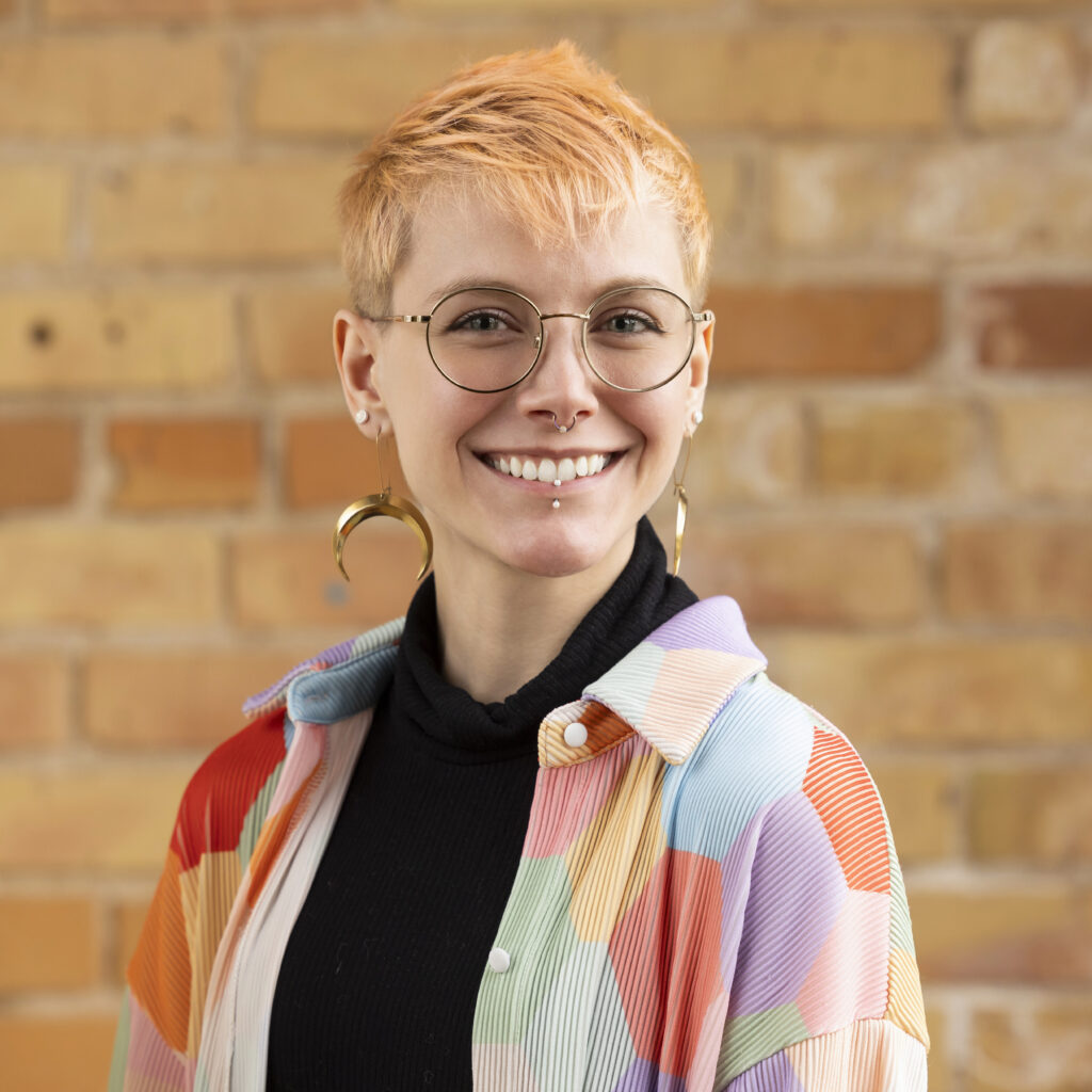 Headshot of a smiling person of light skin tone, with light orange hair and glasses, wearing a colorful rainbow top and large moon earrings.