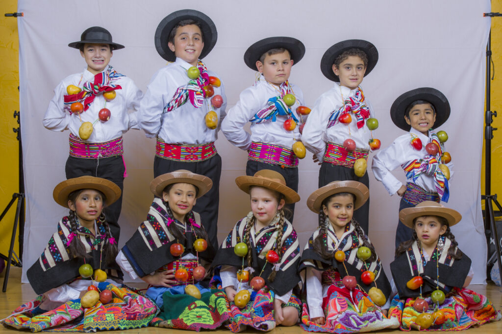 Ten young children, five boys and five girls, wearing traditional Peruvian dance outfits and fruit necklace. The girls wear an embroidered skirt and a blanket on their back and fauna and boys wear black pants and a blanket on their back.