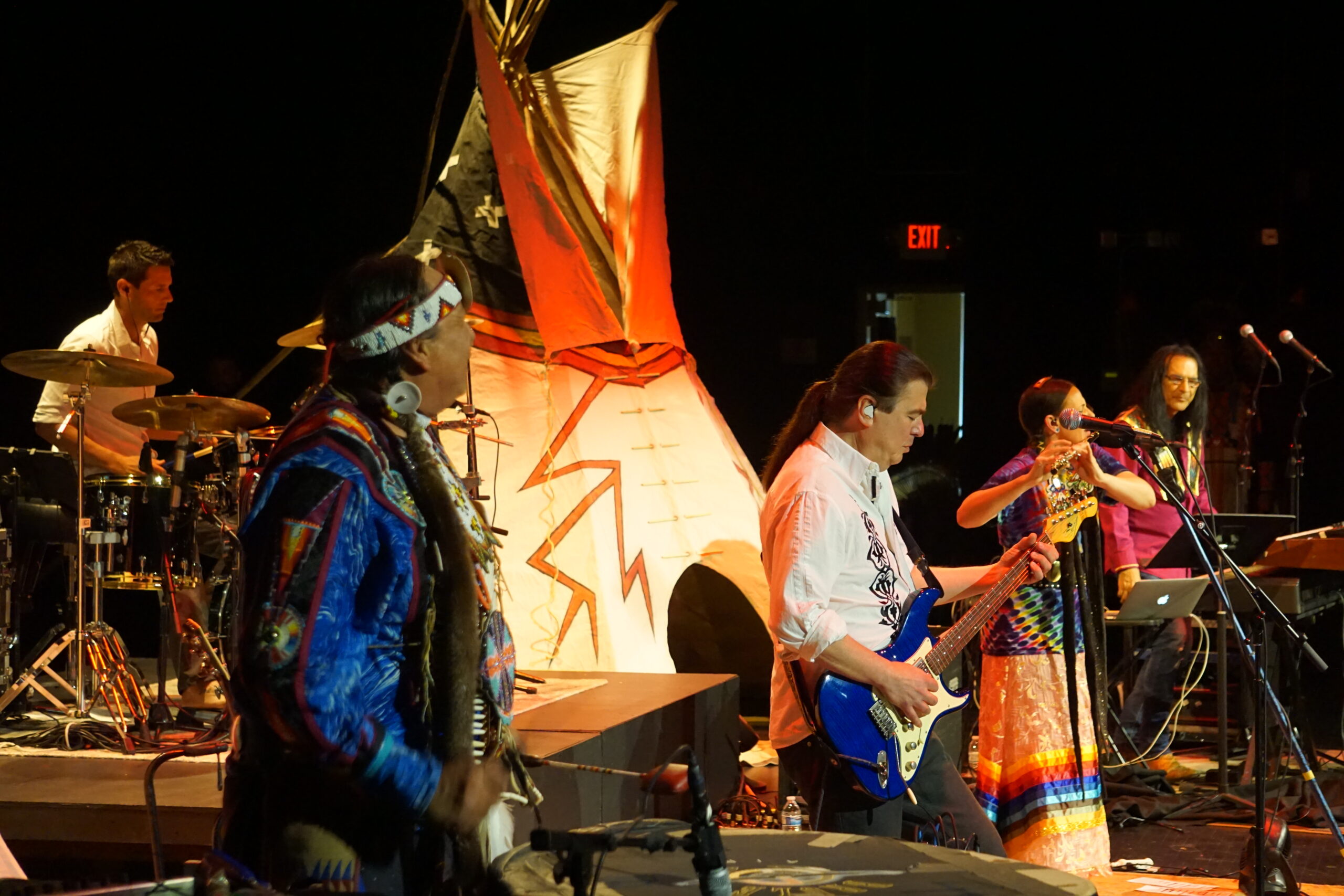 A band on stage, consisting of a drummer, traditional drummer, guitarist, flautist, and keyboardist, some of whom wear traditional regalia, with a tipi set up on stage with them.