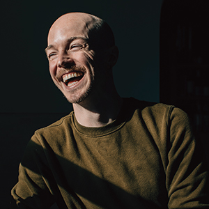 A bald person with a brown mustache laughs with their eyes closed into a singular light source against a black background.