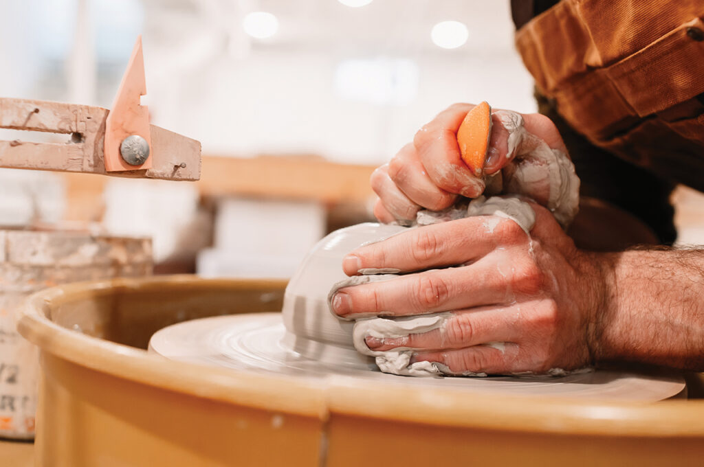 A pair of white hands cup a ball of clay being spun on a wheel.