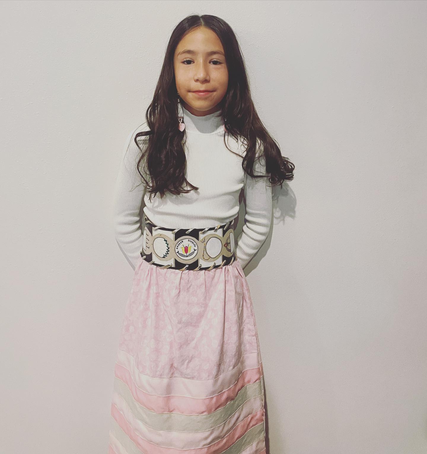 A young person with long dark hair wearing a white turtleneck and a light pink skirt.