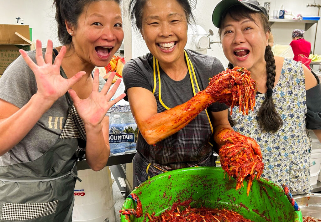 Three smiling women, one holding handfuls of bright red kimchi, stand around a green bucket of kimchi.