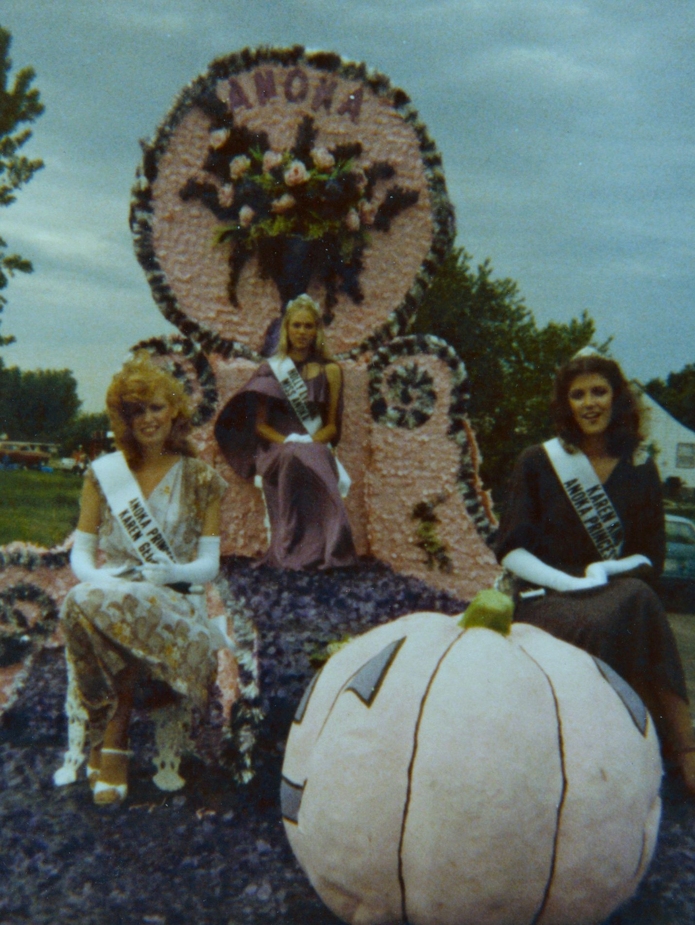 Three people smile on a parade float next to a large pumpkin.