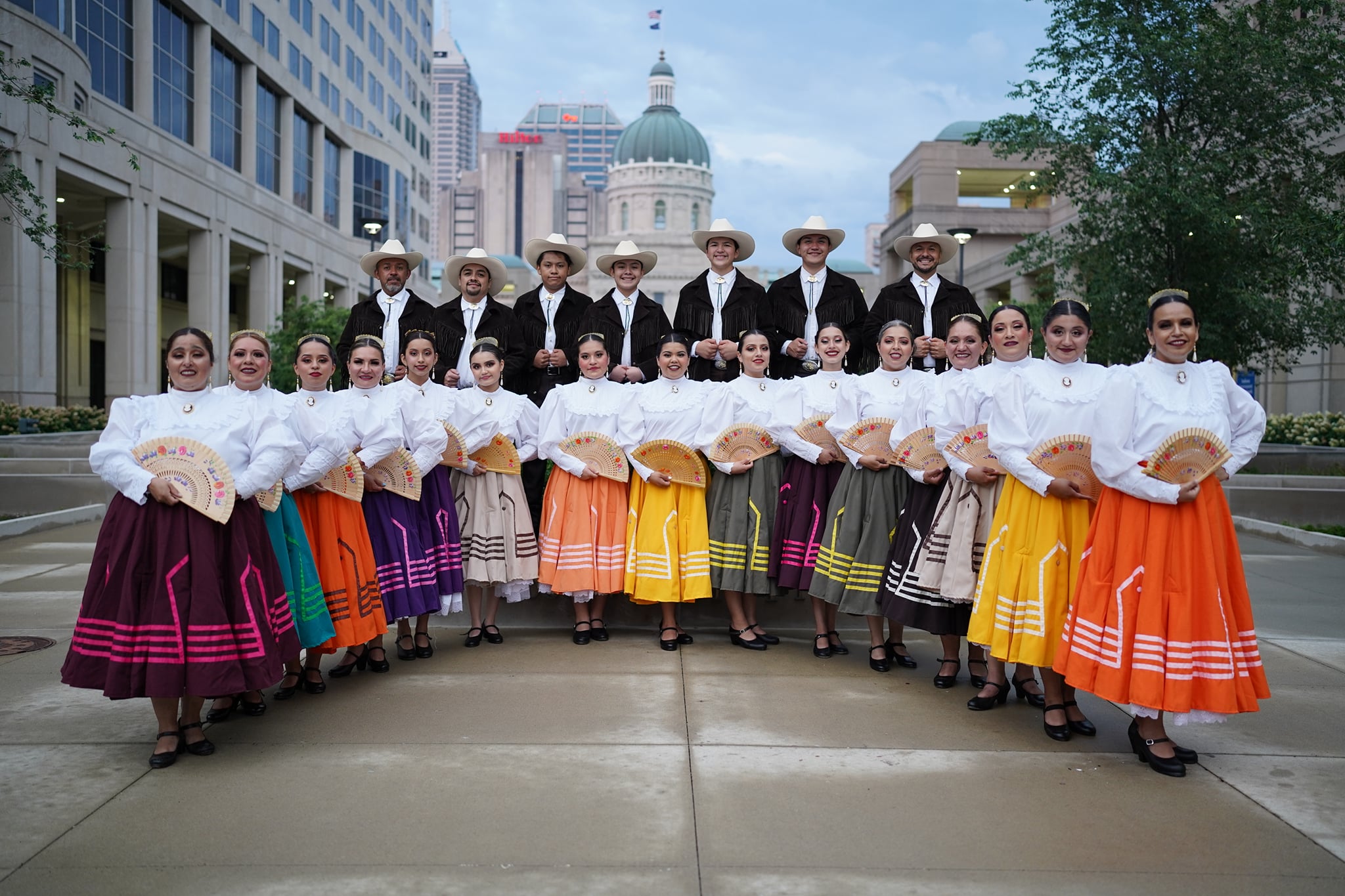 Performers pose and stand in two rows. The dancers in front are wearing colorful traditional skirts with white shirts. The people in the back row are wearing frilled jackets and white hats.