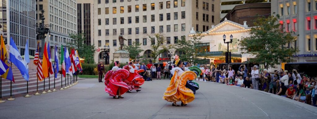 Dancers wearing colorful skirts perform outdoors in front of a crowd. Behind them is a row of flags on flagpoles.