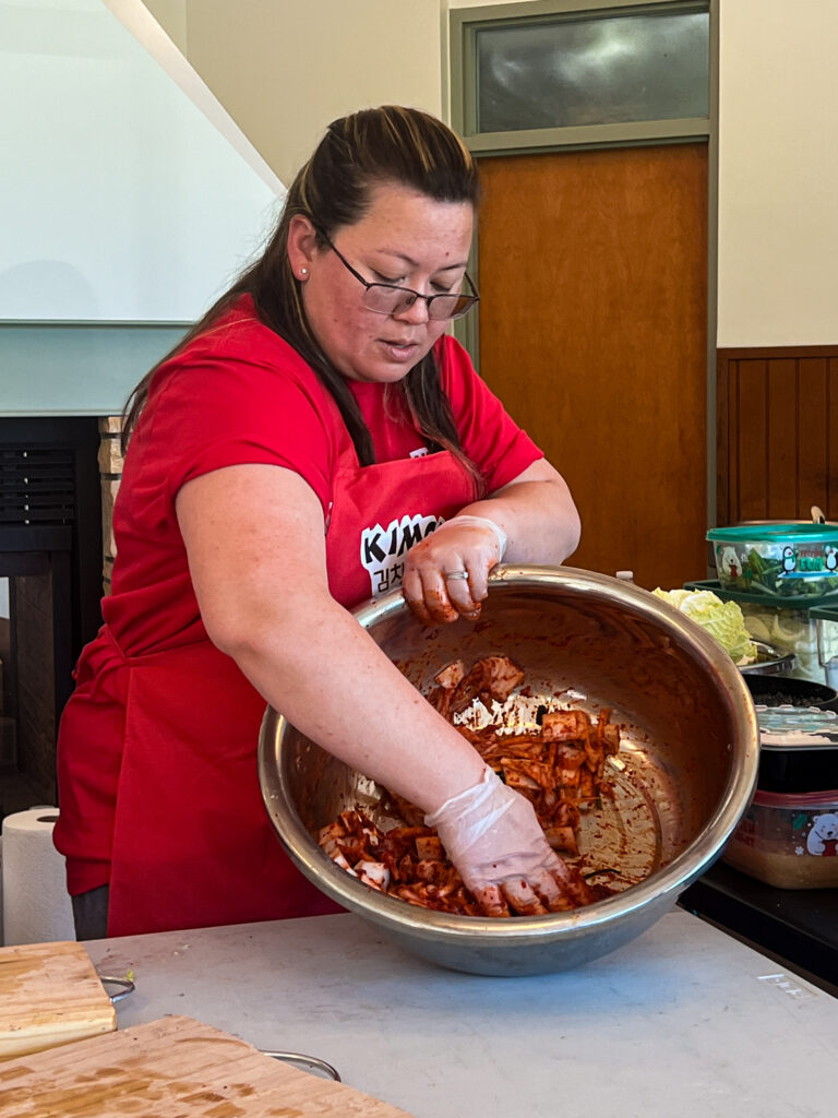 A person with long brown hair wearing glasses hand-mixing a large metal bowl of kimchi.