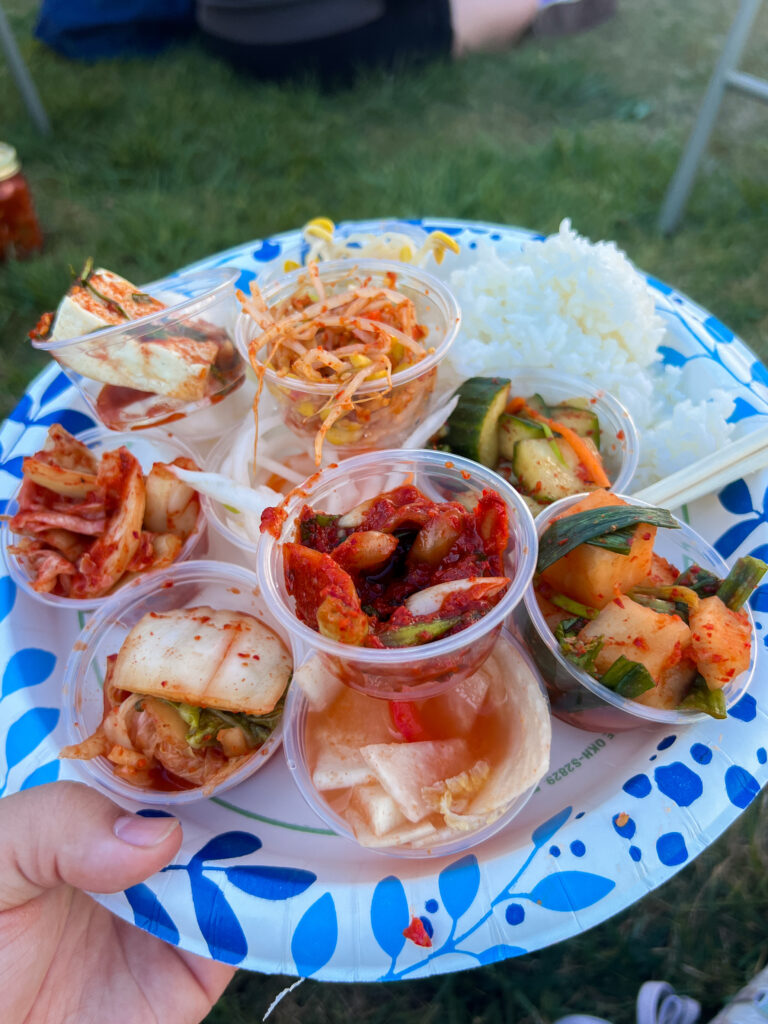 A paper plate stacked high with plastic ramekin cups filled with various varieties of kimchi and a lump of rice.