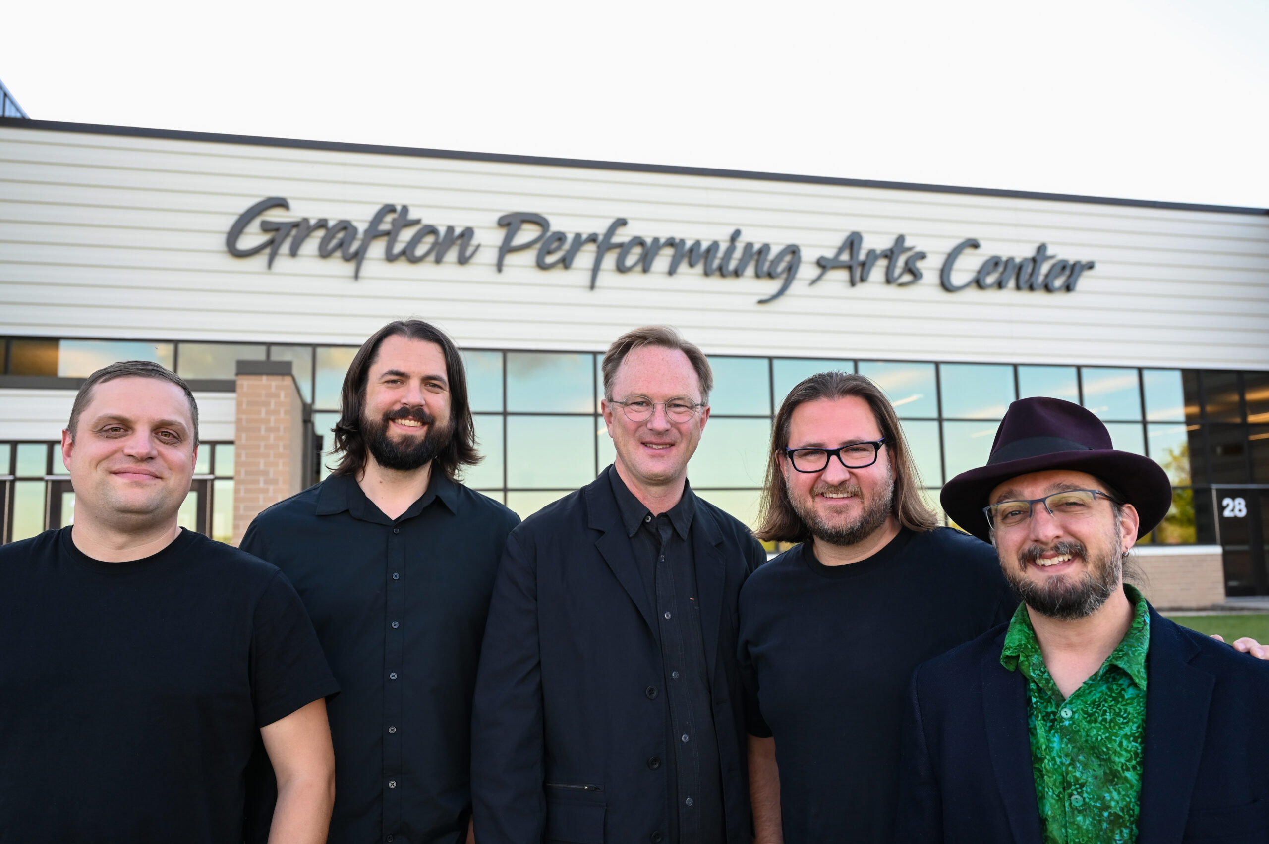 A group of five men in concert black pose for a photo in front of a building whose sign reads Grafton Performing Arts Center