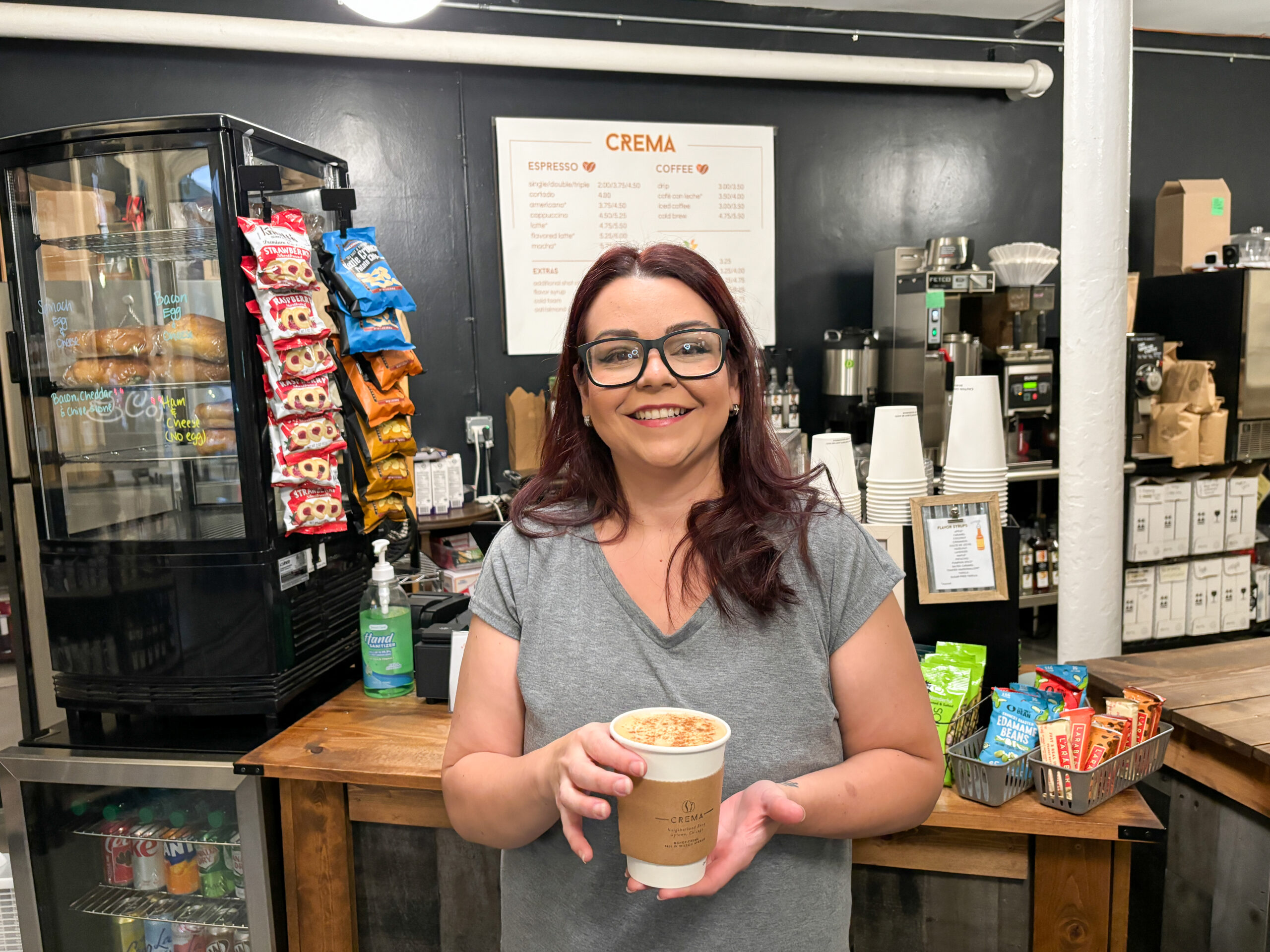 A person with medium skin tone and dark hair smiling and holding a cup of coffee. They are wearing a grey v-neck tshirt and dark-rimmed glasses.