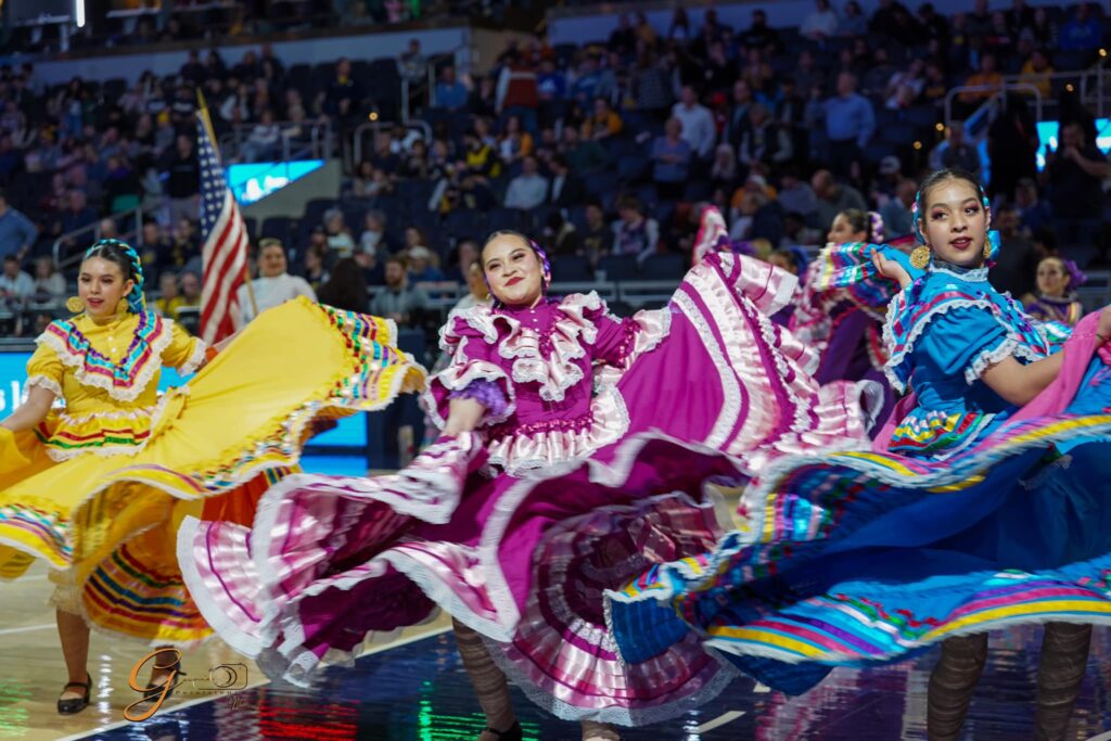 Dancers wearing colorful, traditional dresses as they perform at a basketball arena.