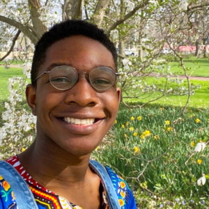 Headshot of smiling person with dark skin tone wearing overalls and a multicolored short in front of a garden.