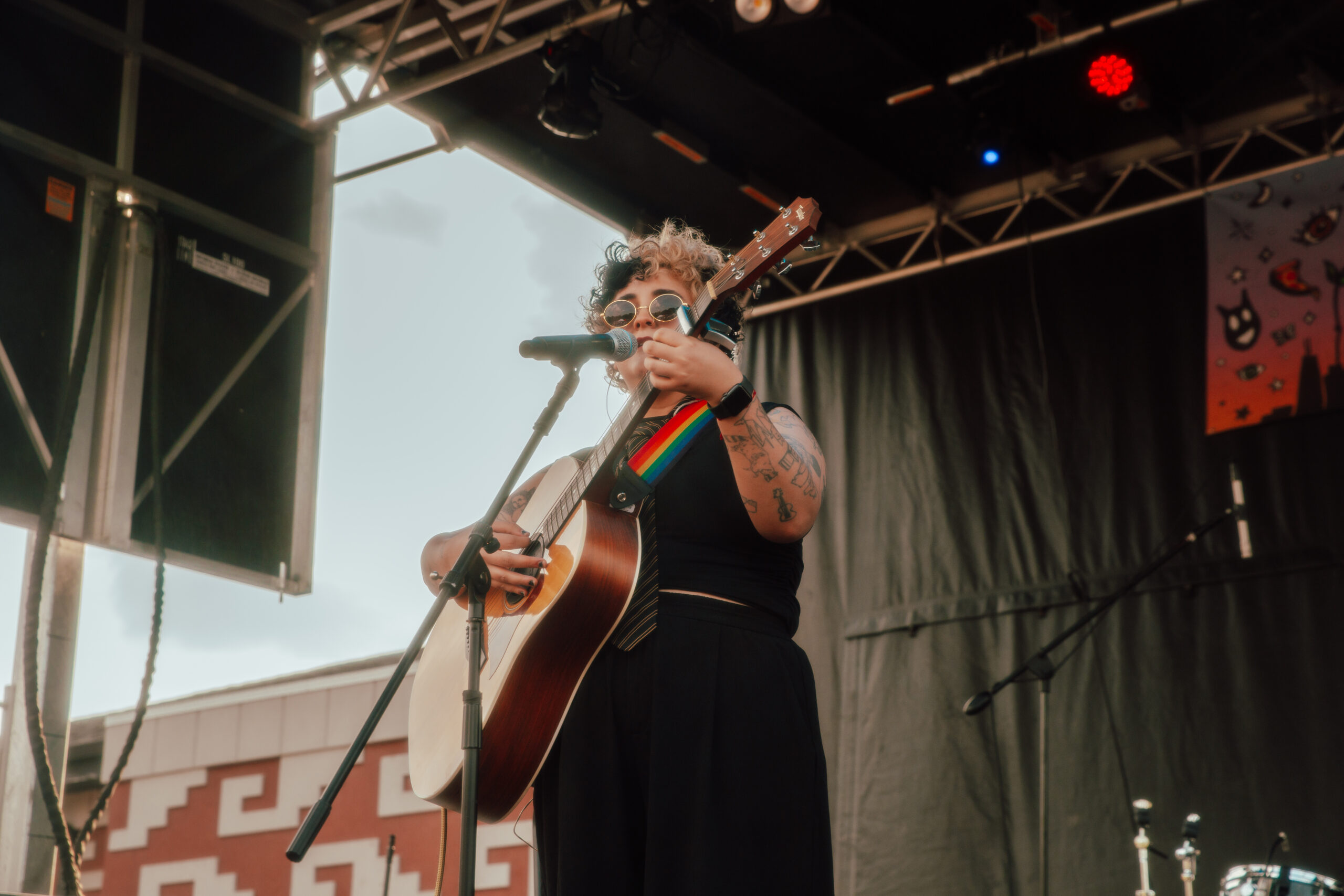 A person of medium skin tone standing and singing into a microphone. They are playing an acoustic guitar.