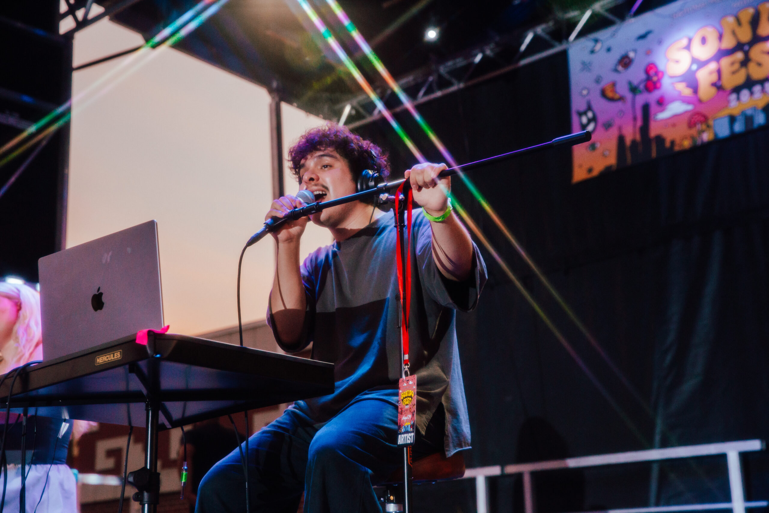 A person of medium skin tone and short curly hair performs on stage. They are sitting on a chair and singing into a microphone.