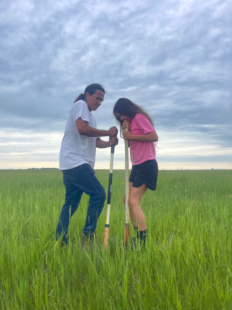 Two people holding shovel-like tools to dig the ground. The ground has knee-high green grass.