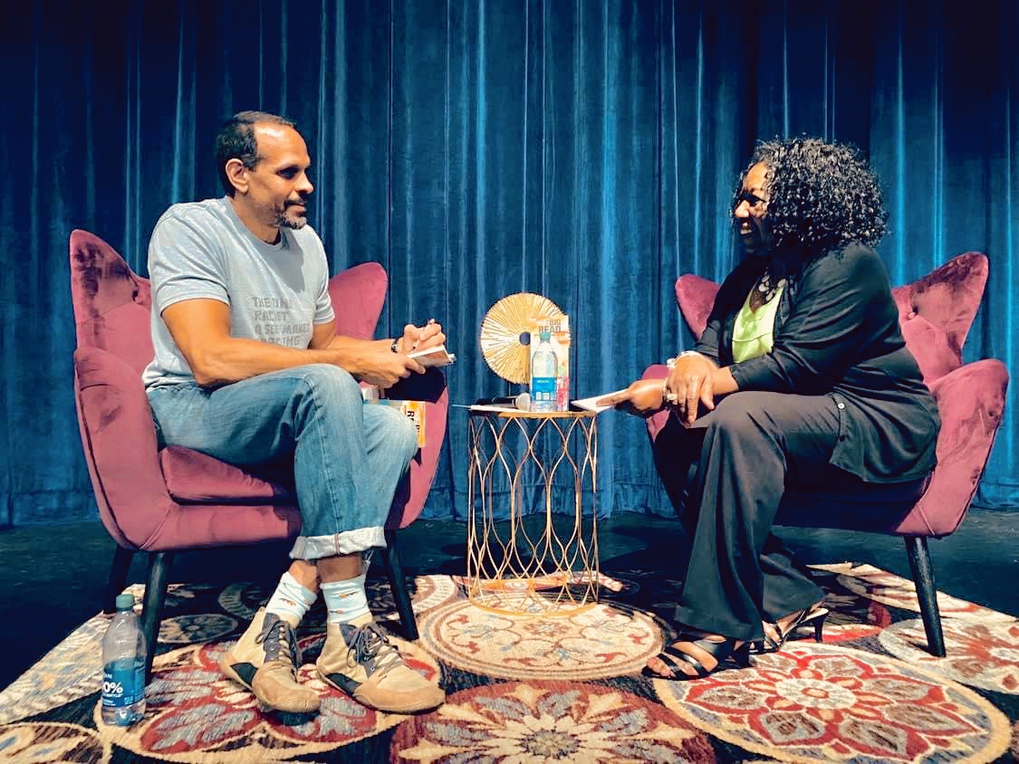 Two people of medium dark skin tone sit in pink wingback chairs against a blue velvet background.