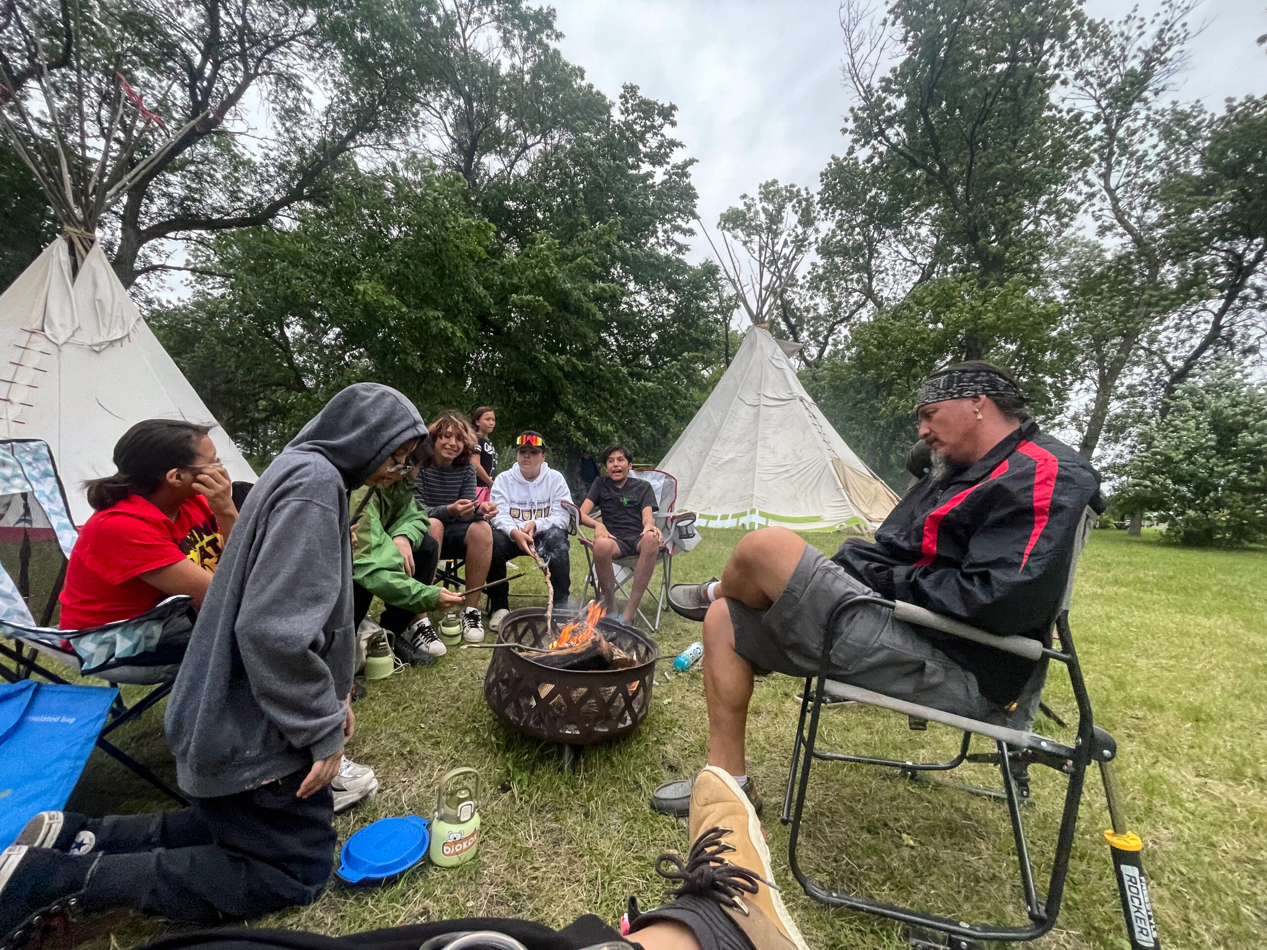 A group of youth sitting around a fire outdoors as they look to the adult sitting in front of them.