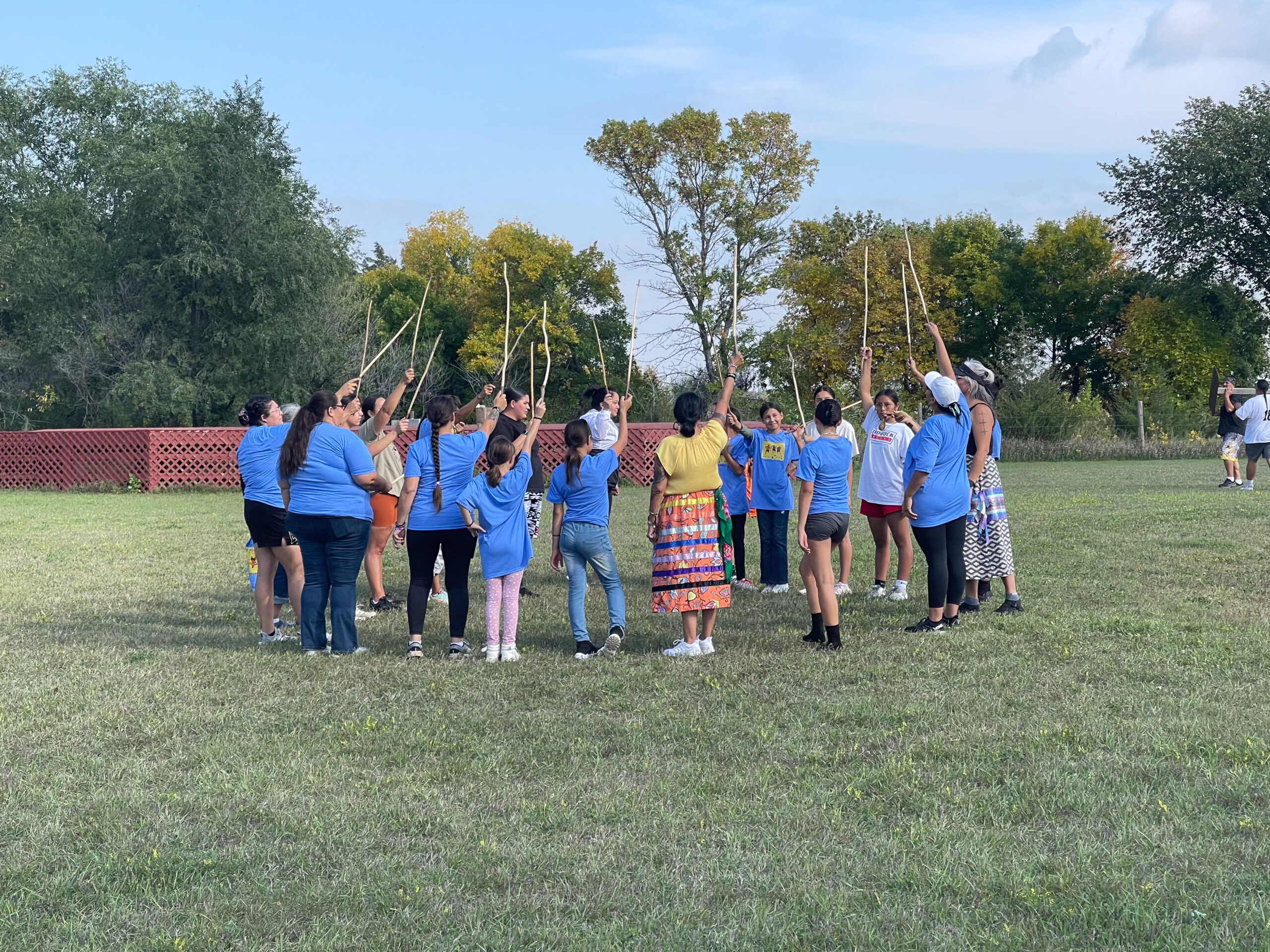 A group of adults and children standing in a circle on a field. They are holding sticks up in the air. Some of them are wearing bright blue tshirts.