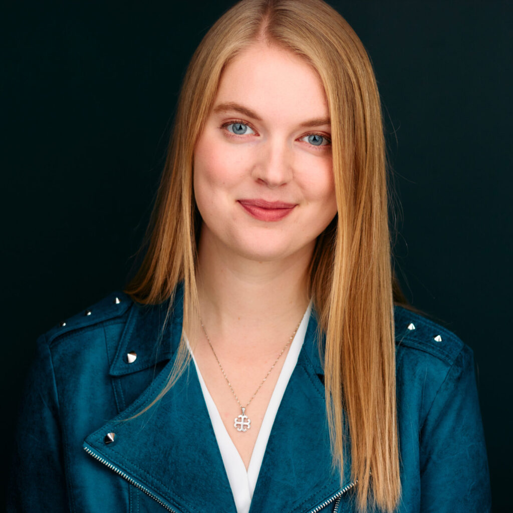 Headshot of a smiling person with light skin tone and long blonde hair, wearing a turquoise blazer and a silver necklace.