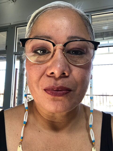 A person a medium skin tone wearing a dark half-frame glasses. They have their hair tied back and are wearing long earrings.