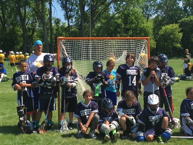 A group of children wearing dark purple hockey uniforms standing in front of a goal with one adult in the background.