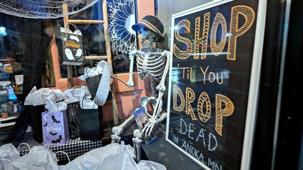 A sign reads "Shop Till You Drop Dead in Anoka MN" next to a skeleton decoration inside a store.