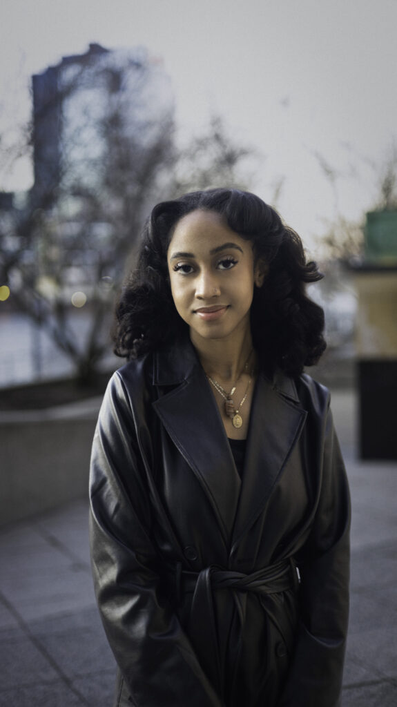 Headshot of a person with shoulder-length curly black hair, medium skin tone, and a leather peacoat.