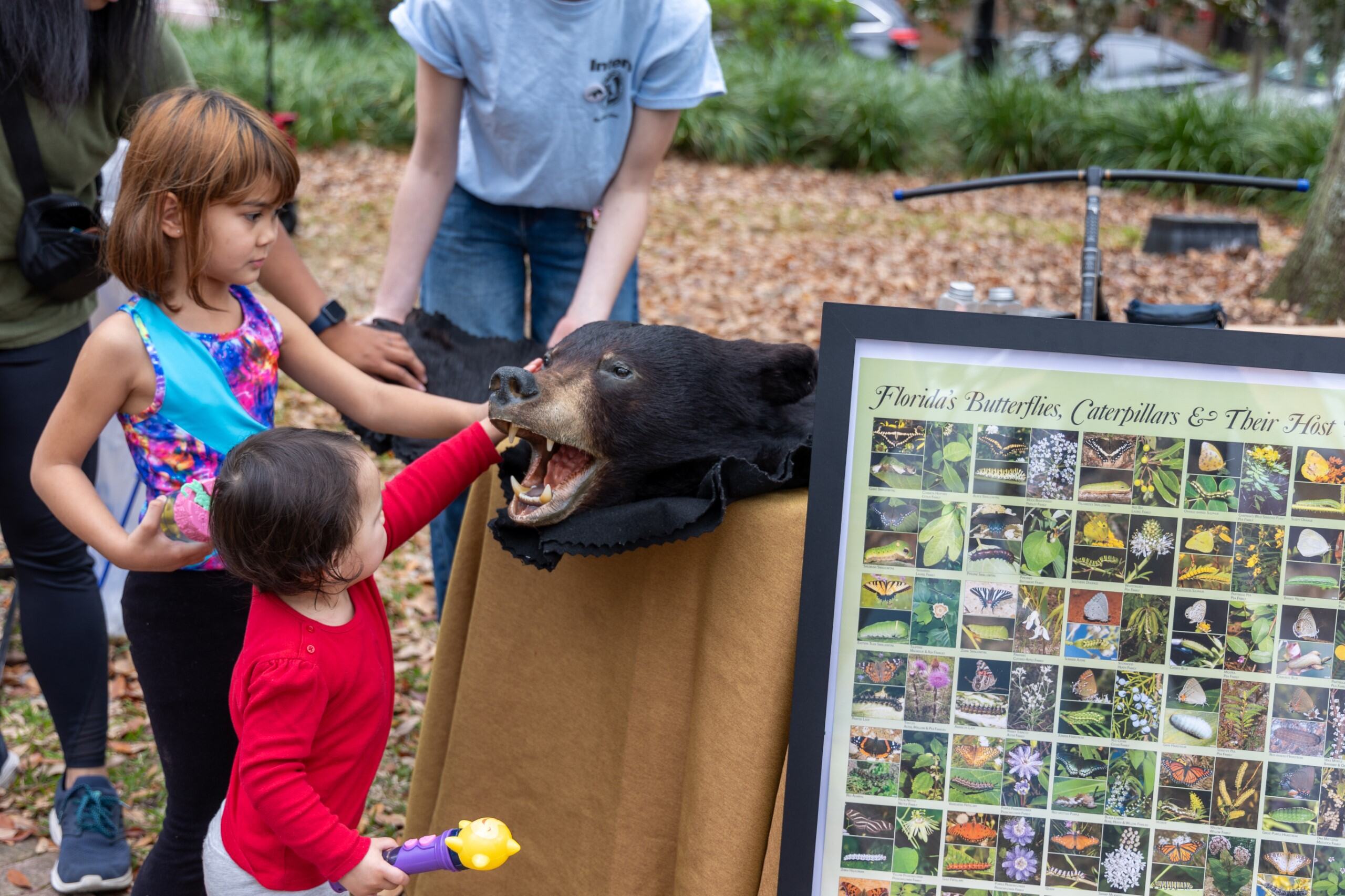 Two young children, one in a red shirt and the other in a multi-colored tank top, interact with the pelt of a black bear.