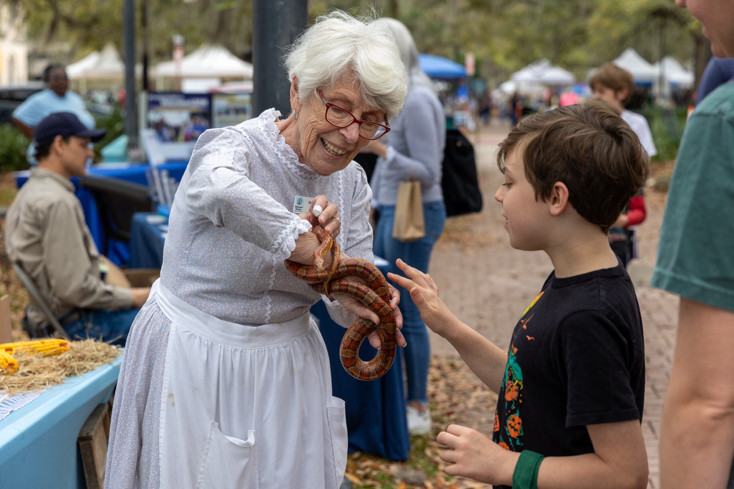 An older woman with white hair, red glasses, and a white dress, shows a young boy in a black shirt a snake. The snake is wrapped around her wrist and is red and brown and black.