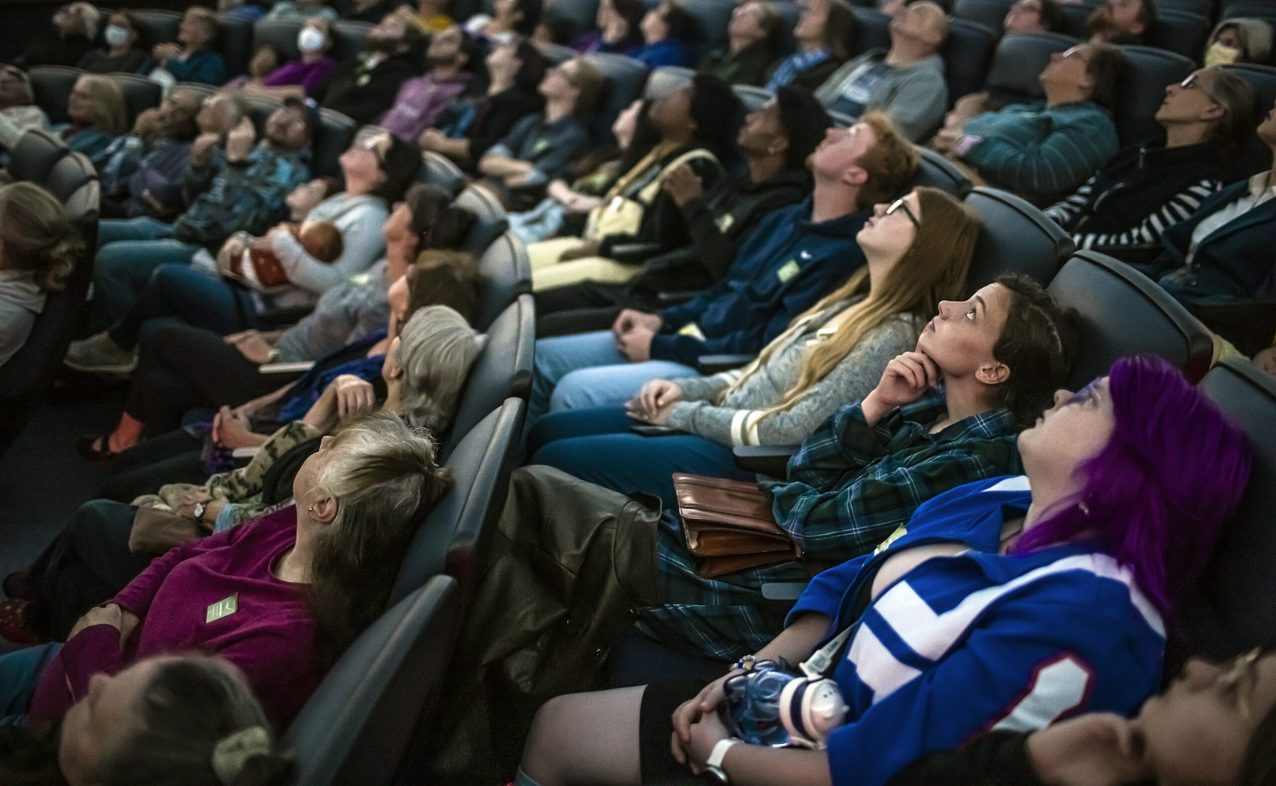 A theater full of people sitting in auditorium seats and looking upward.