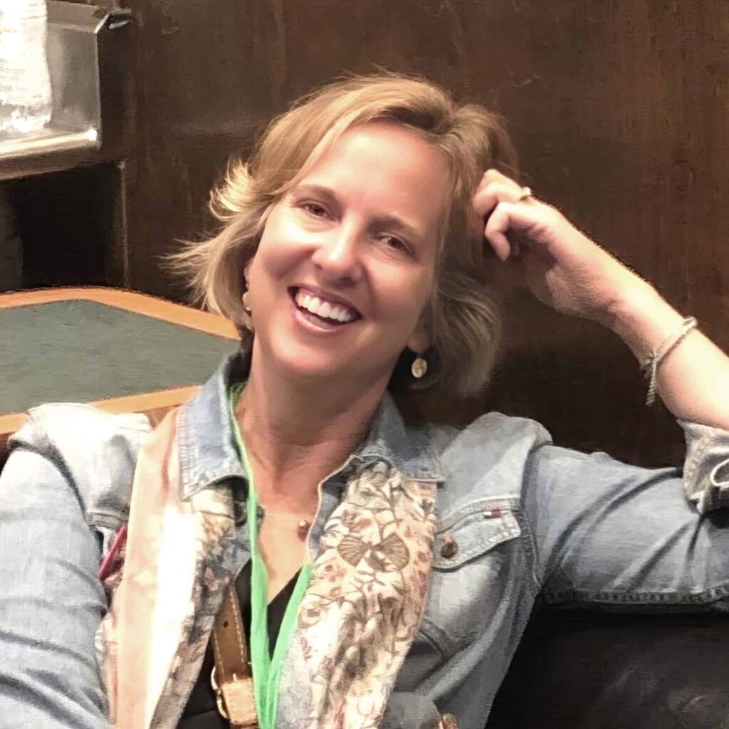 Headshot of a smiling woman with light skin tone and shorter blondish hair, wearing a denim jacket and scarf, and leaning casually with her elbow bent and hand resting on her head.