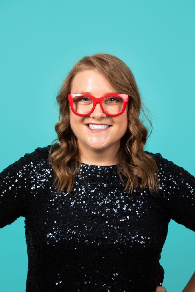 Headshot of a smiling woman, with light skin and wavy auburn hair, wearing a black sequin shirt and red glasses.