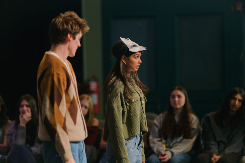 Two actors, one in a fox mask, stand on a stage in front of audience members.