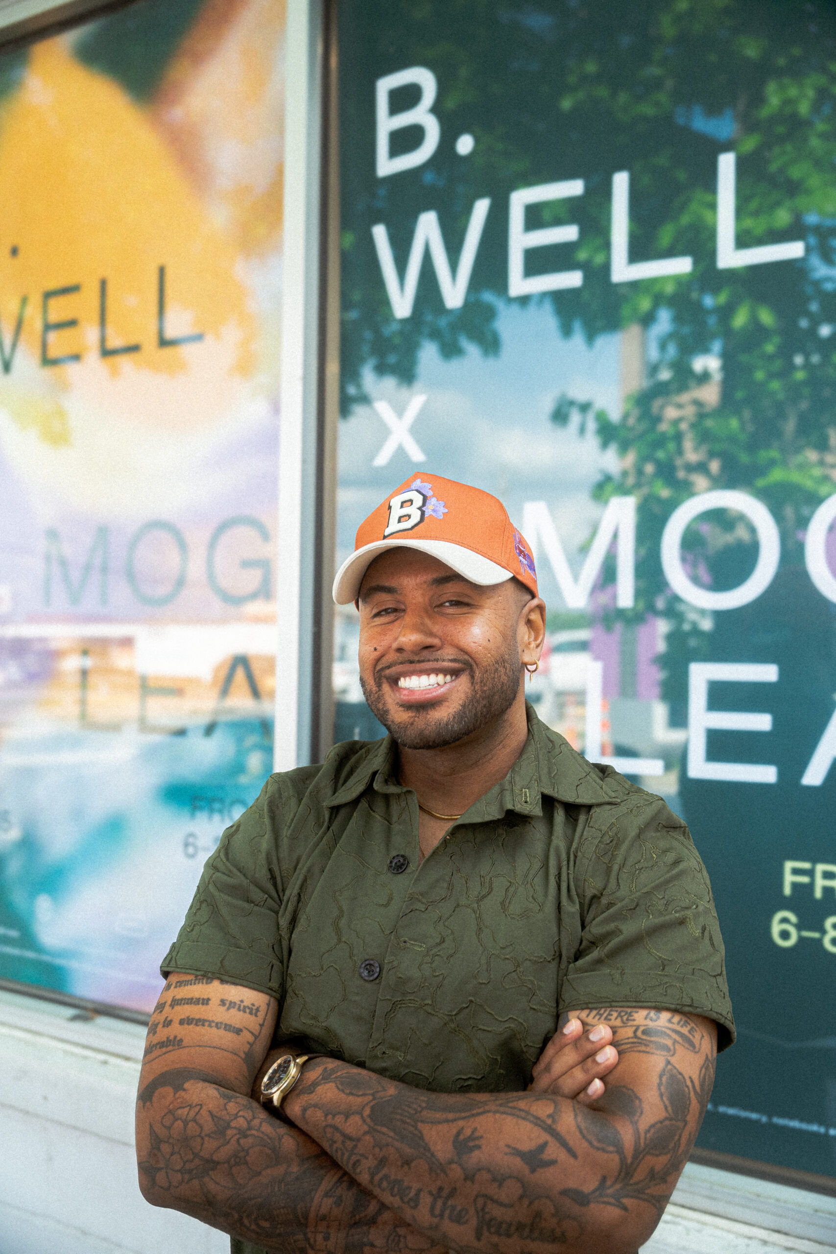 A man with dark skin and tattoos in a green shirt and orange hat stands in front of a sign that reads B. Well