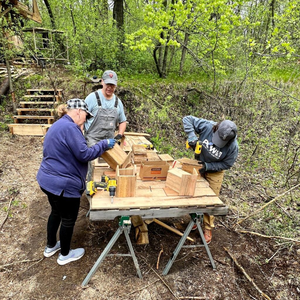 A group of three people build birdhouses on a table in the woods