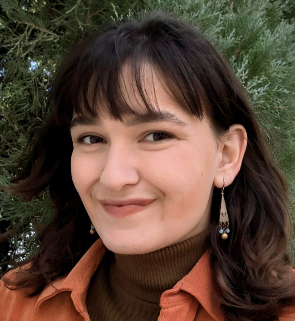 Headshot of a smiling person with light skin tone and dark brown hair, wearing an orange jacket.