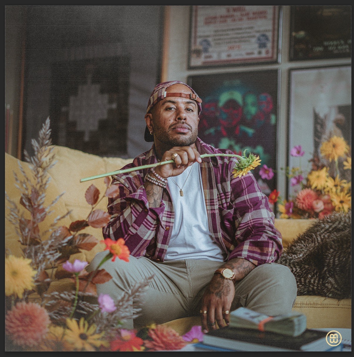 A man with dark skin in a plaid shirt and red hat surrounded by fake flowers.