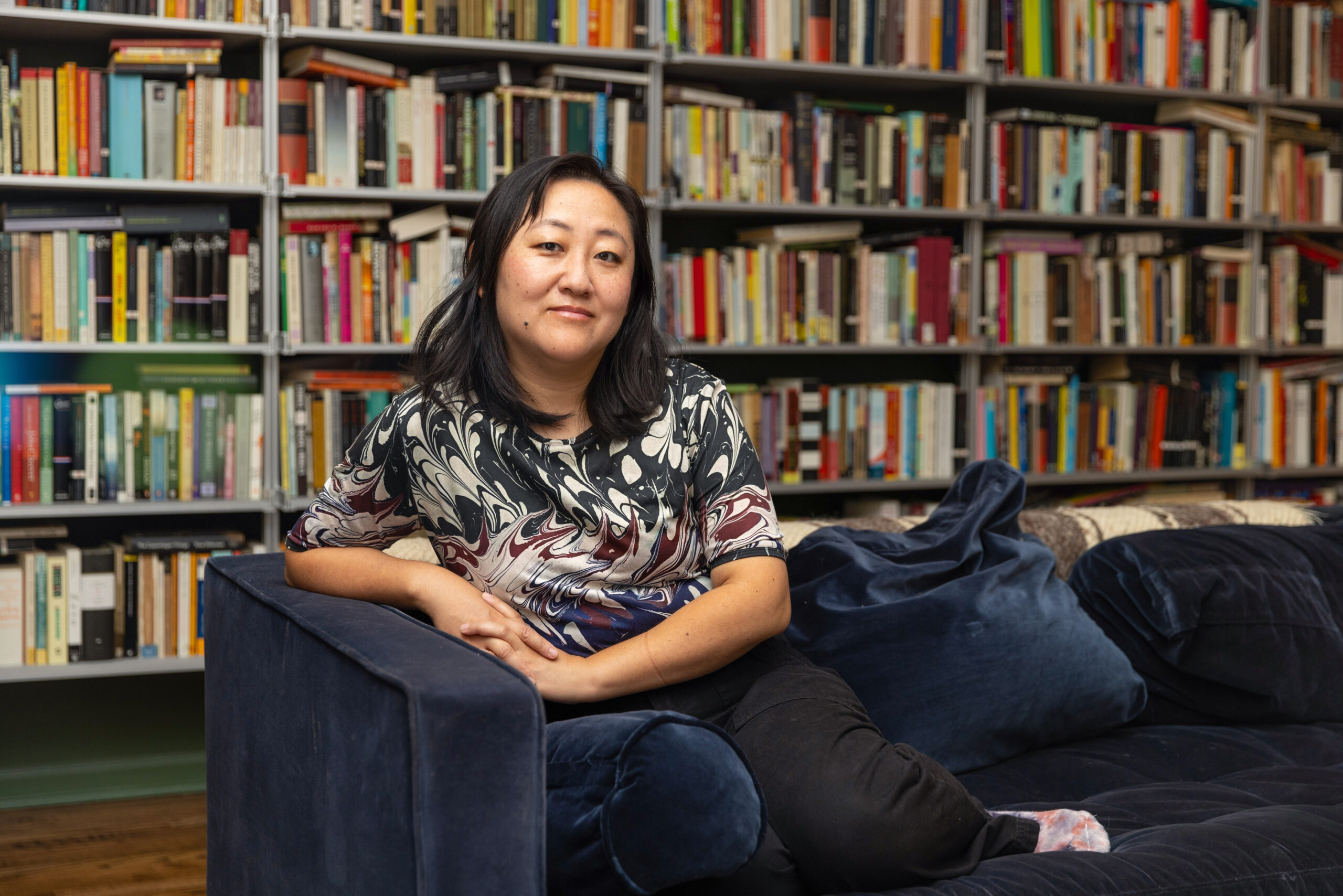 A person of medium skin tone and shoulder-length dark hair sits on a navy blue sofa. Behind them is a large wall-mounted shelf filled with books.