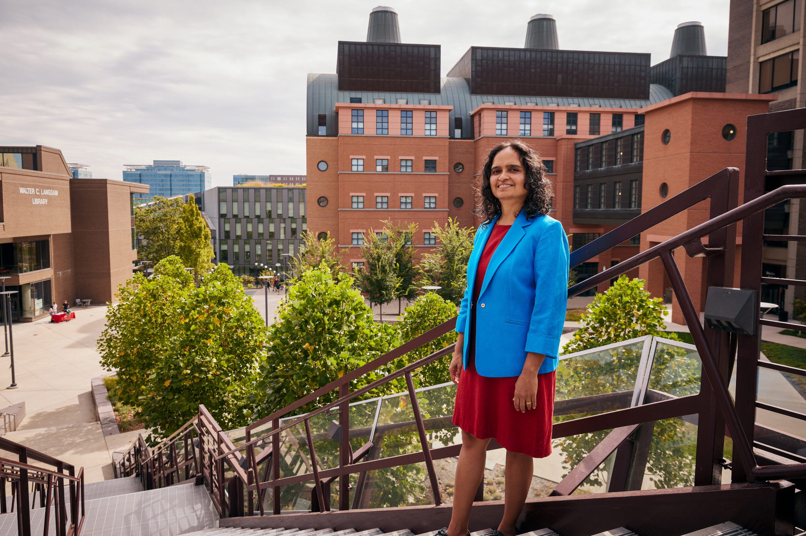 A person of medium skin tone with dark, shoulder-length wavy hair stands at the top of a large outdoor staircase. Behind them are tall brick buildings.