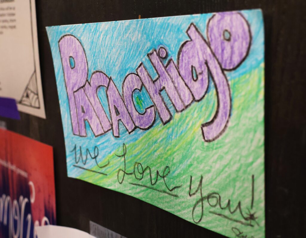 A handdrawn paper sign with bold purple letters that spells "parachigo," and the phrase "we love you!" written under it.