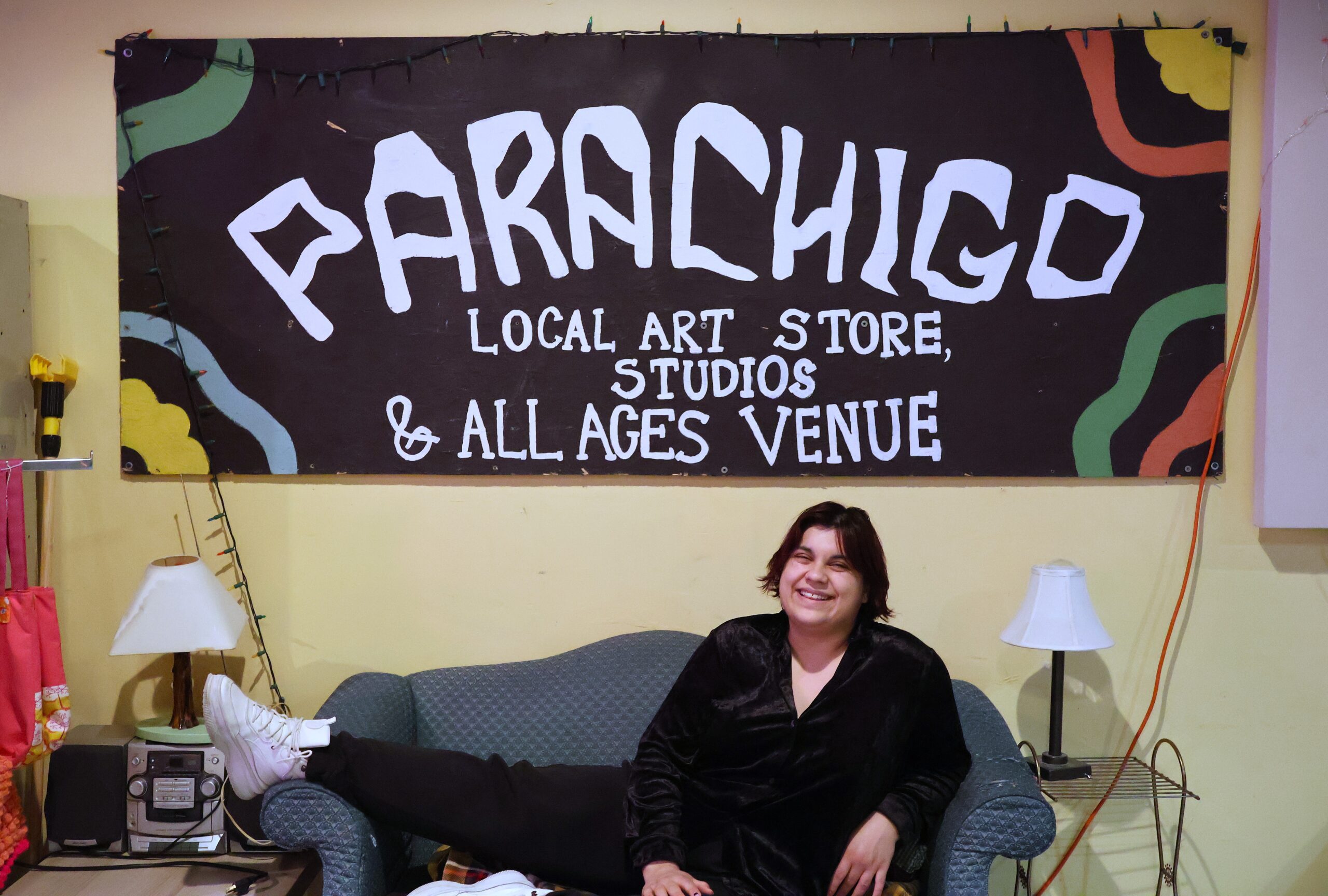 A person wearing black smiles and poses with their leg up on a couch under a large sign.