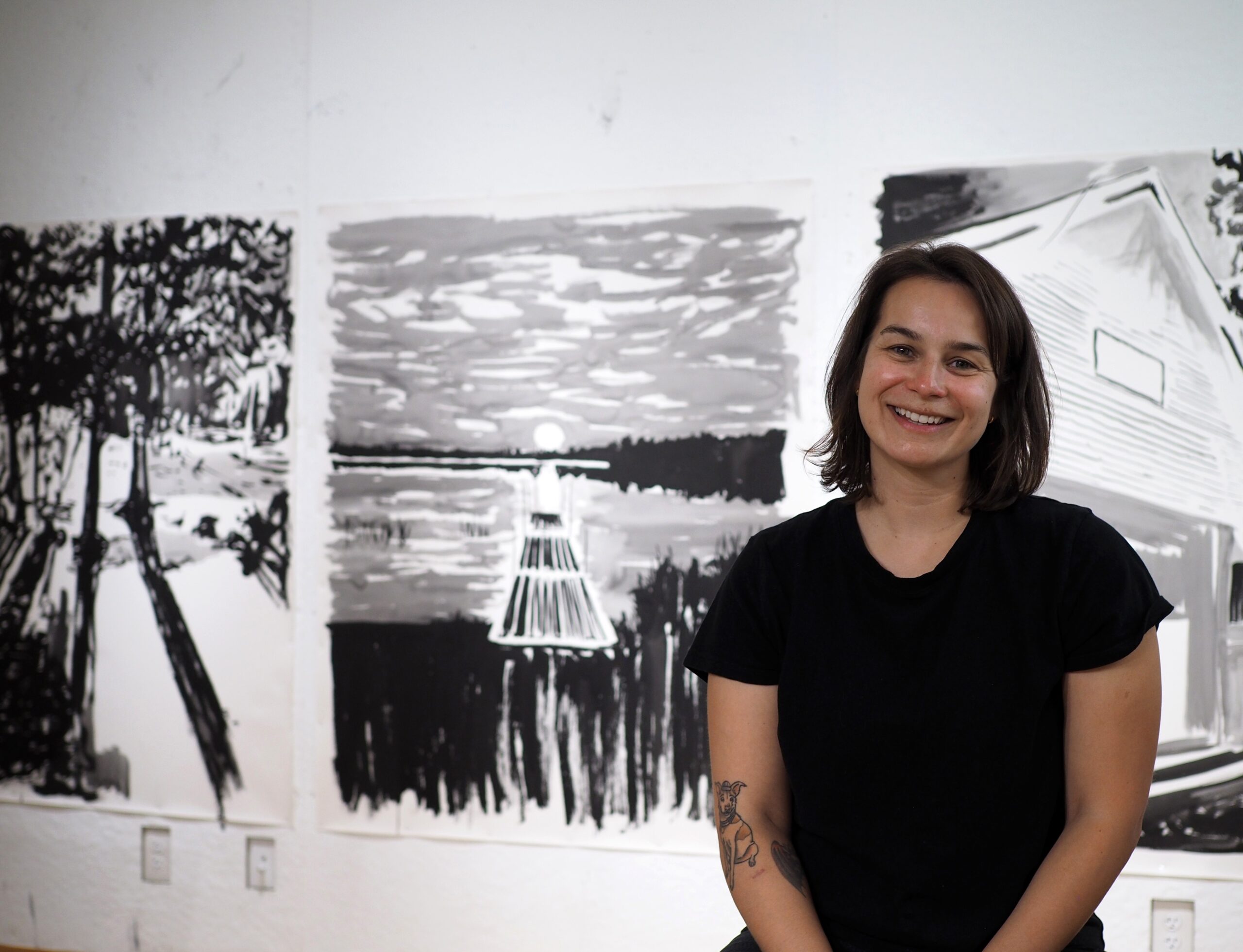 A woman wearing black sits and smiles in front of three art pieces.