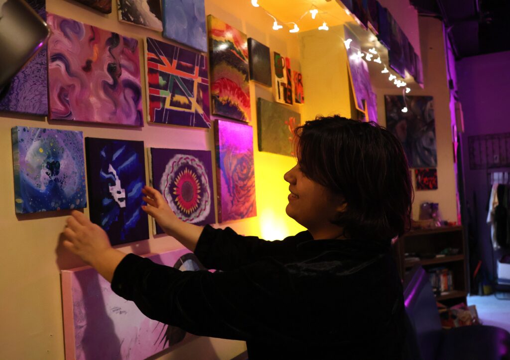 A person with dark hair and dark colored clothing with their arms raised as they adjust a painting on the wall in front of them.