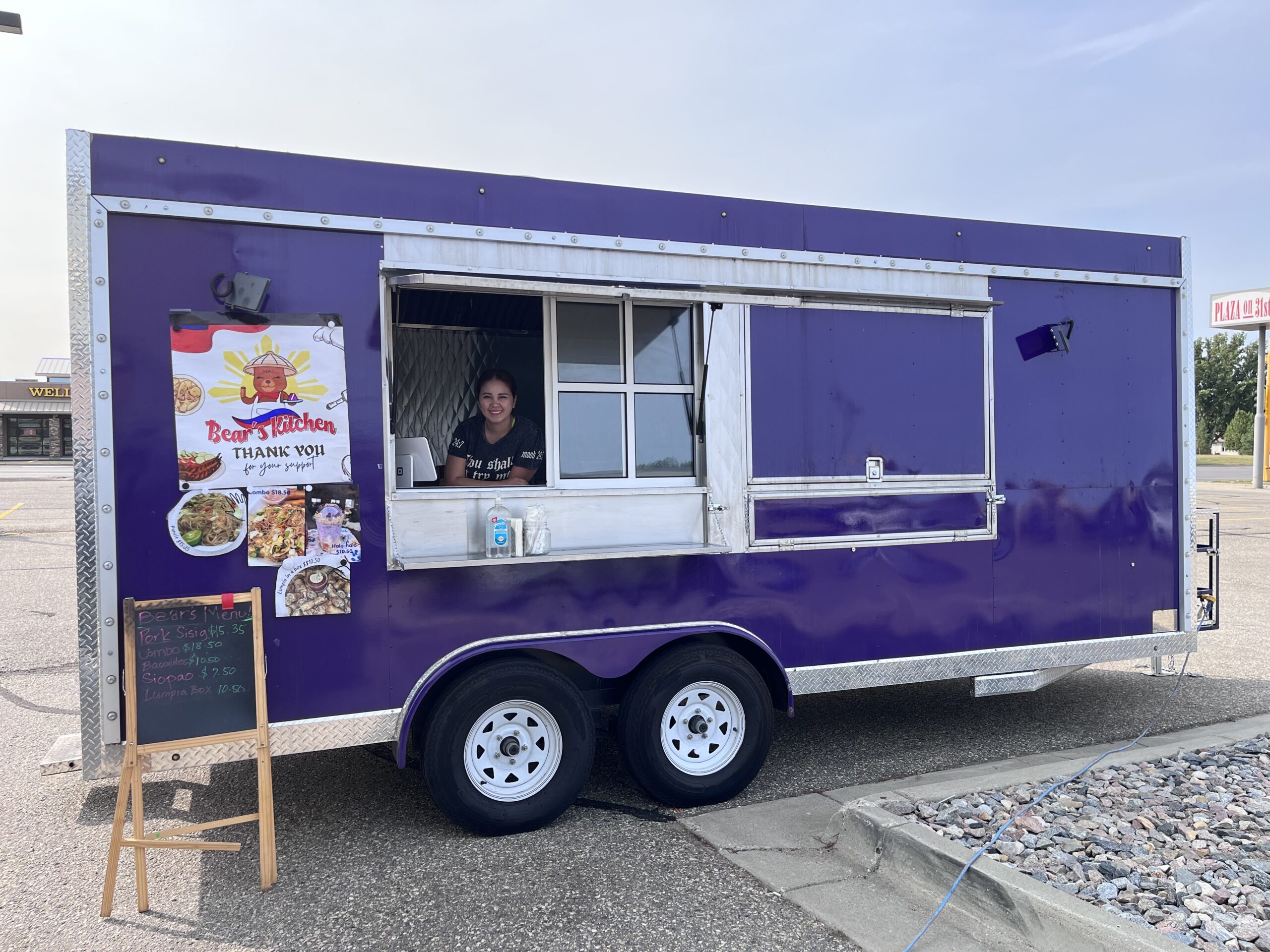 A purple food truck with a person looking out of the window. It is parked in parking lot.