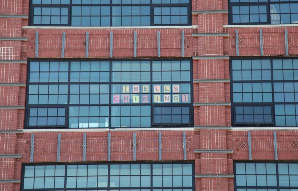 A brick building with large windows. One of the windows has letters printed on colorful paper. The letters spell '[bells chiming]'