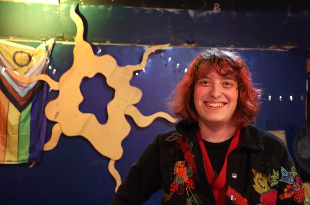 A person of light skin tone smiling. They have colored hair and are wearing a dark shirt with floral embroidery on it.